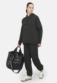 Sudadera con capucha negra y bolsillo frontal tipo canguro, combinada con pantalones de chándal a juego. Sosteniendo una bolsa de hombro de puffer negra brillante con asas.