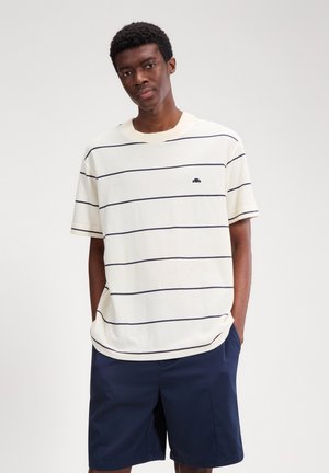 Junger Mann trägt ein weißes Kurzarmhemd mit dünnen horizontalen schwarzen Streifen und marineblaue Shorts, die Hände in den Taschen.