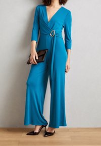 Combinaison enteal à décolleté en V profond, manches trois-quarts et accent en boucle dorée. Associée à une pochette noire et des talons hauts.