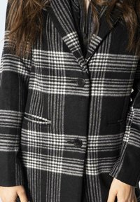 Černobílý kostkovaný kabát s klopovým límcem, dvěma předními kapsami a zapínáním na dvojaké knoflíky. Vyrobeno z materiálu s texturou podobnou vlně.