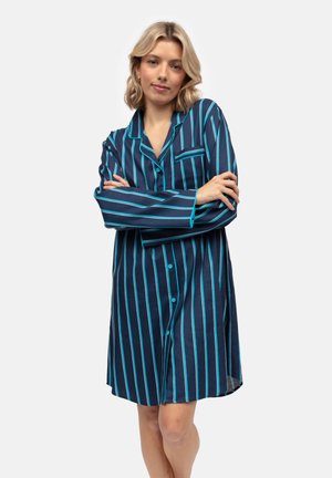 Giovane donna in piedi con le braccia incrociate, che indossa una camicia da notte blu navy a lunghezza ginocchio con strisce verticali turchesi e bianche.