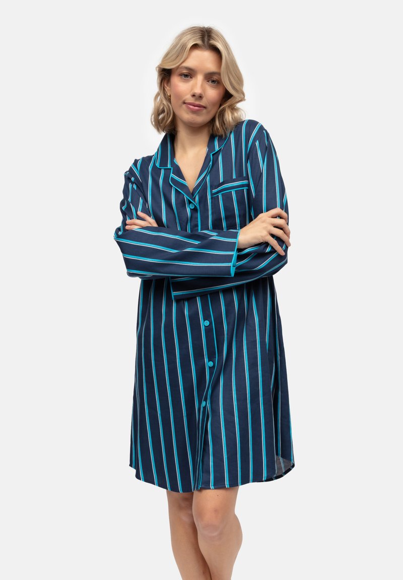 Młoda kobieta stoi z założonymi rękami, ubrana w nocną koszulę do kolan w granatowym kolorze z pionowymi turkusowymi i białymi paskami.