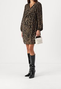 Žena v dlouhém rukávu s hnědým a černým zvířecím vzorem, černými kozačkami nad kolena a držící malou bílou kabelku.