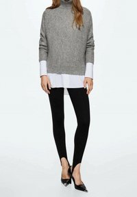 Grauer, strukturierter Rollkragenpullover über einem weißen Hemd, kombiniert mit schwarzen Leggings und schwarzen spitzen Absatzschuhen mit silbernen Akzenten.