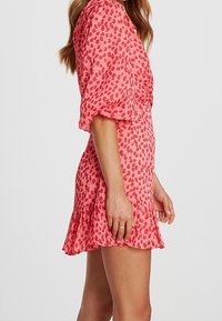 Růžové šaty s krátkým rukávem a volánkovým lemem, se skvrnitým vzorem v tmavší růžové. Vyrobeno z lehké strukturované tkaniny.