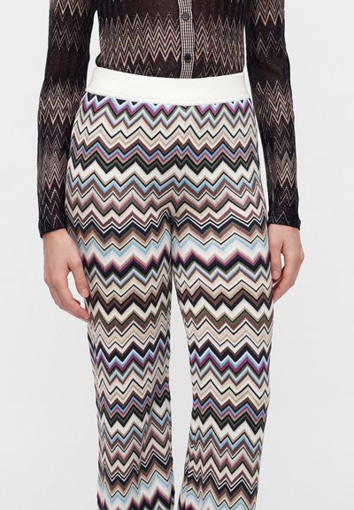 Vue de la taille jusqu'au milieu de la cuisse d'une personne portant un pantalon taille haute avec un motif zigzag multicolore et un haut noir transparent à manches longues avec un design géométrique.
