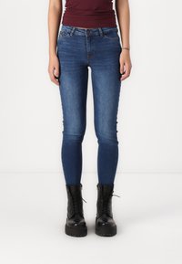 Μπλε τζιν skinny παντελόνι με μεσαία μέση, πέντε τσέπες και λεπτομέρειες ραφής, συνδυασμένο με μαύρες μπότες με κορδόνια.