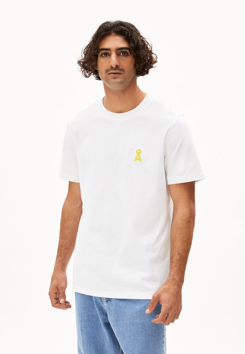 Witte katoenen t-shirt met een ronde hals, korte mouwen en een kleine gele afbeelding op de linkerkant van de borst. Draagt het met lichtblauwe denim jeans.