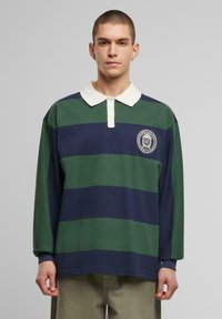 Long-sleeve rugby shirt featuring green and navy horizontal stripes, a white collar, and a circular logo on the chest. Soft cotton fabric.
