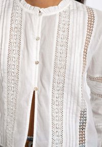 Blusa blanca con cuello con volantes, cierre central de botones y paneles de encaje verticales. Presenta una textura de tela suave y un delicado detalle de trabajo de calado.