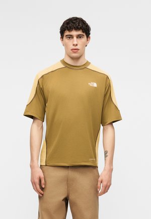 Man met kort krullend haar, gekleed in een bruin-beige kortärmelig The North Face-shirt en beige broek, staand tegen een effen achtergrond.