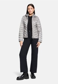 Veste matelassée gris clair avec capuche, fermeture éclair frontale et poches latérales, associée à un pantalon noir et des bottes noires épaisses. Texture lisse.