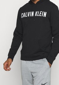 Hombre vistiendo una sudadera negra de Calvin Klein y pantalones de chándal gris claro de Nike, de pie con una mano en el bolsillo y la otra mano relajada.