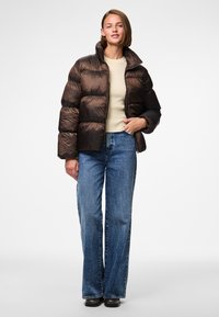 Brun puffjacka med hög krage, lättviktigt material och quiltad struktur, bärs över en cremefärgad topp och blå vida jeans.