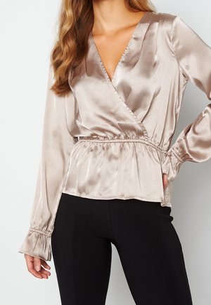 Femme portant un chemisier portefeuille en satin couleur champagne avec taille et poignets élastiques, associé à un pantalon noir ajusté, sur un fond clair.