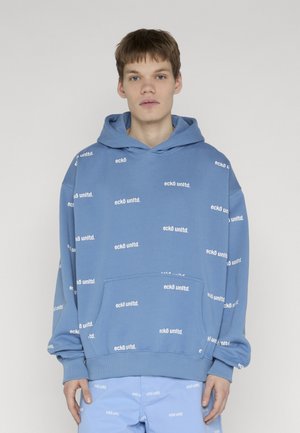 Junger Mann mit kurzen braunen Haaren, der einen blauen übergroßen Hoodie und passende Shorts trägt, bedeckt mit einem weißen "eckō unltd." Schriftzugmuster.