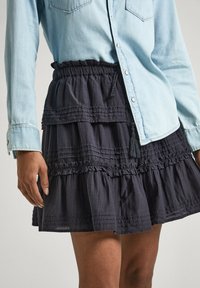 Osoba oblečená v černé mini sukni s volánky a světle modré denimu košili na knoflíky, ruka odpočívající po boku.