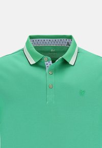 Grön pikétröja med texturerat tyg, tre-knapps knäppning och kontrasterande vita krage-accenter. Har en liten broderad logotyp på bröstet.