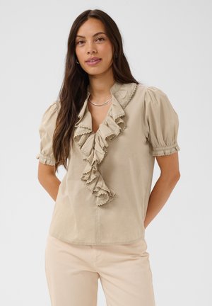 Femme aux longs cheveux bruns portant une blouse beige à volants avec des manches bouffantes et un pantalon clair, debout devant un fond uni clair.