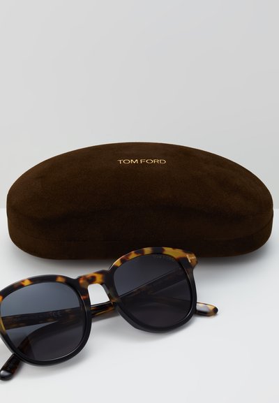 Tortoiseshell zonnebril met donkere lenzen, voorzien van een uitgesneden ontwerp, naast een bruin suède hoesje met "TOM FORD" in goud gedrukt.