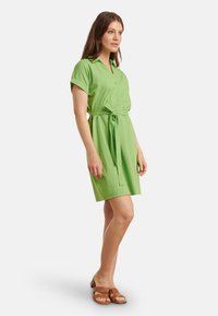 Grünes Kurzarmkleid mit einem kragenlosen Ausschnitt und einem gebundenen Taillenband. Hergestellt aus leichtem Stoff, hat es eine lockere Passform und eine Länge über dem Knie.
