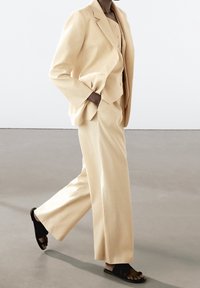 Beżowy garnitur na miarę z szerokimi spodniami i jednołopatkową marynarką, w połączeniu z czarnymi płaskimi sandałami. Gładka tekstura materiału, strukturalny fason.