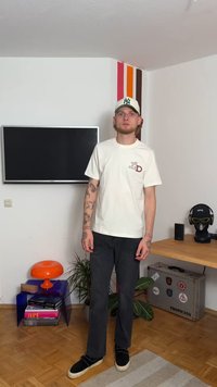 Gaiši krēmkrāsas t-krekls ar bordo grafiku, apvienots ar melniem taisnajiem džinsiem un melnām slīdējošām kurpēm. Iekštelpu vide ar dekoratīviem elementiem.