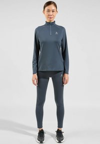 Donkergrijze langesleeve top met een half-zip kraag, gecombineerd met bijpassende slim-fit leggings, beide gemaakt van een gladde, rekbare stof. Zwarte sportschoenen.