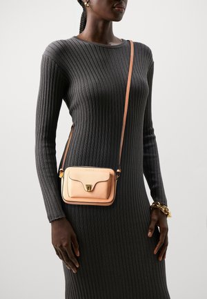 Torebka na ramię z jasnopomarańczowej skóry o wyraźnej, strukturalnej formie, z złotymi okuciami i zapięciem na klapę. Noszona na dopasowanej szarej prążkowanej sukience.