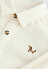 Chemise couleur crème en tissu texturé, avec un bouton marron et un petit design brodé d'un chien teckel sur la poitrine.