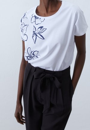 Vit t-shirt med blå blommönster på ena sidan, kombinerad med högmidjade svarta byxor som har en rosett vid midjan.