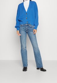 Blå, överstor stickad cardigan med ribbat mönster och v-ringning, prydd med tre stora knappar. Kombinerad med utsvängda blå jeans och svarta stövlar.