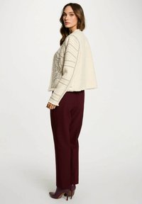 Donna con lunghi capelli castani, che indossa un maglione crema a fantasia, pantaloni a gamba larga bordeaux e stivali con tacco alto bordeaux abbinati, in piedi di profilo.