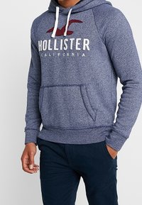 Muž v modré mikině Hollister s bílými šňůrkami a tmavě modrých kalhotách, stojící s jednou rukou v kapse a druhou ruku uvolněnou.