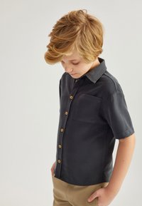 Camisa de botones de manga corta en gris oscuro con botones de madera, un bolsillo en el pecho y textura suave, combinada con pantalones beis claros.