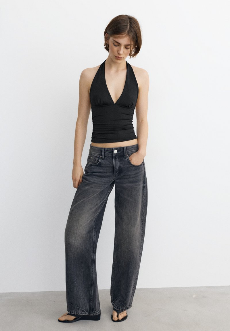 Jeune femme debout portant un haut dos nu noir, un jean noir délavé à jambes larges, et des tongs noires sur un fond blanc uni.
