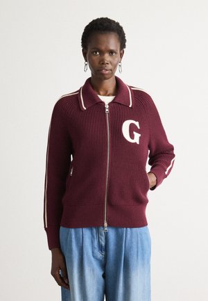 Woman wearing a maroon zip-up sweater with white stripes on sleeves and a large white "G" on the chest, paired with loose blue jeans.