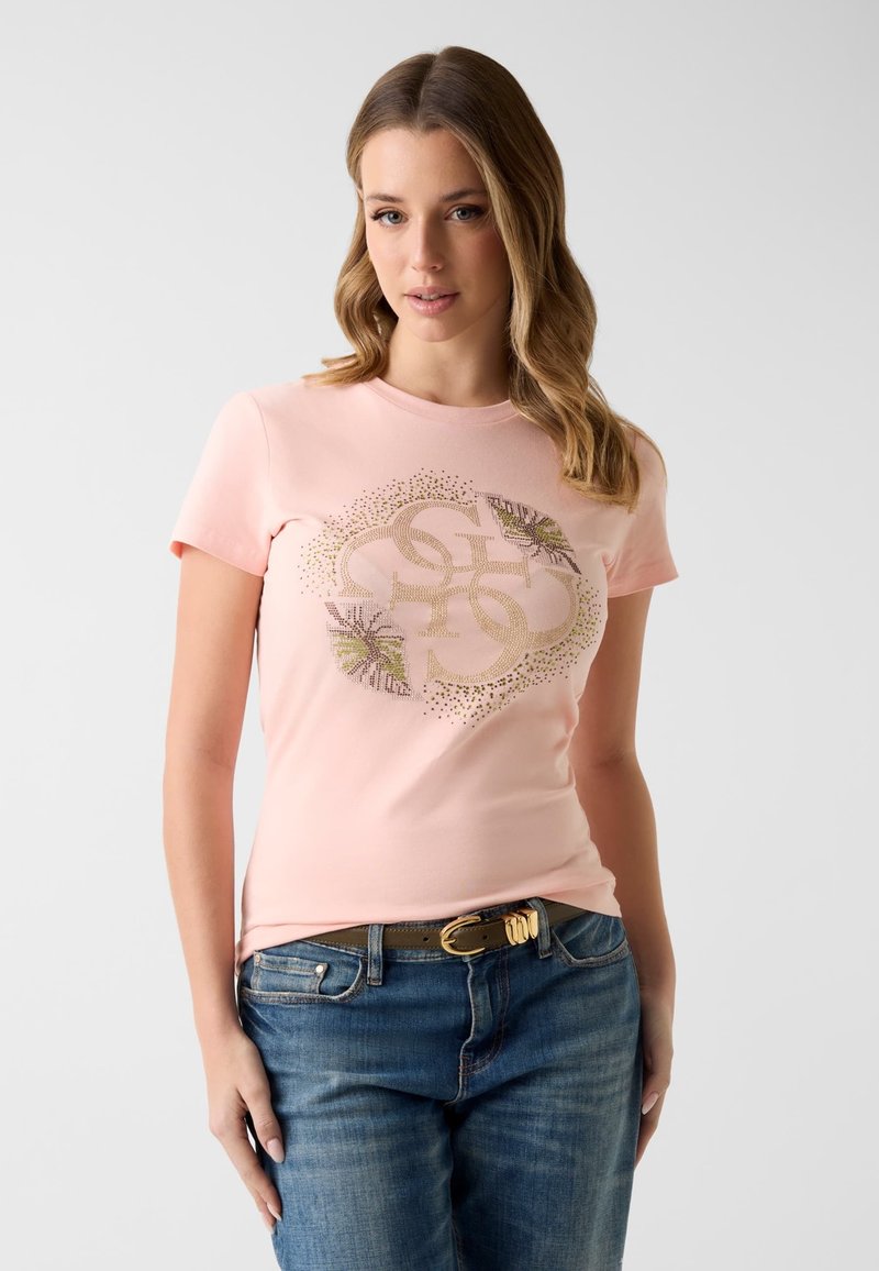 Žena s volnými vlnitými vlasy nosí světle růžové tričko se zlatým dekorativním vzorem a modré džíny s tmavým páskem.