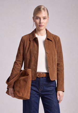 Young woman wearing a brown suede jacket with matching bag, white top, blue jeans, and brown belt, standing against a plain background.