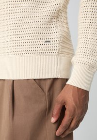 Pull en tricot beige crémeux avec un tissage texturé, des poignets et un ourlet côtelés. Présente une petite étiquette en métal près de la couture latérale. Assorti à un pantalon marron.