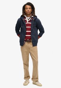Marine blauwe ritshoodie over een bordeauxrode en witte gestreepte polo, gecombineerd met kaki broek en beige sneakers, met een geruit accent op de taille.