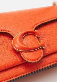 Sac en cuir orange vif à texture granuleuse avec une grande fermeture incurvée bordée d’or sur le rabat avant.