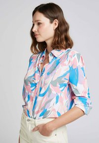 Blouse colorée avec un motif floral abstrait dans des tons de rose, bleu et gris. Design léger, avec des boutons et des manches retroussées.