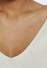 Top en tricot côtelé crème avec un décolleté en V profond, présentant une texture subtile et un matériau doux et léger. Accent sur la clavicule et la zone de la poitrine.