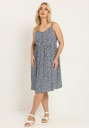 Blond kvinde iført en mørkeblå blomsterprikket, ærmeløs midi-kjole med spaghetti-stropper og hvide kilehælsandaler, stående på en ensfarvet baggrund.