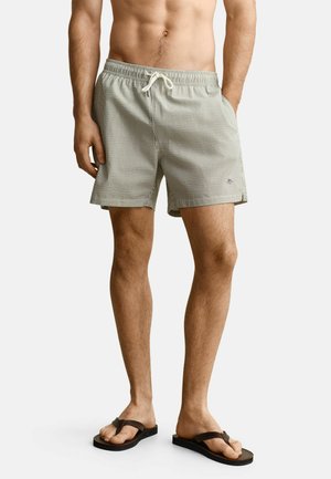 Man draagt lichtgrijze gestreepte zwemshort met trekkoord en donkergroene slippers, staat met één hand in zijn zak.