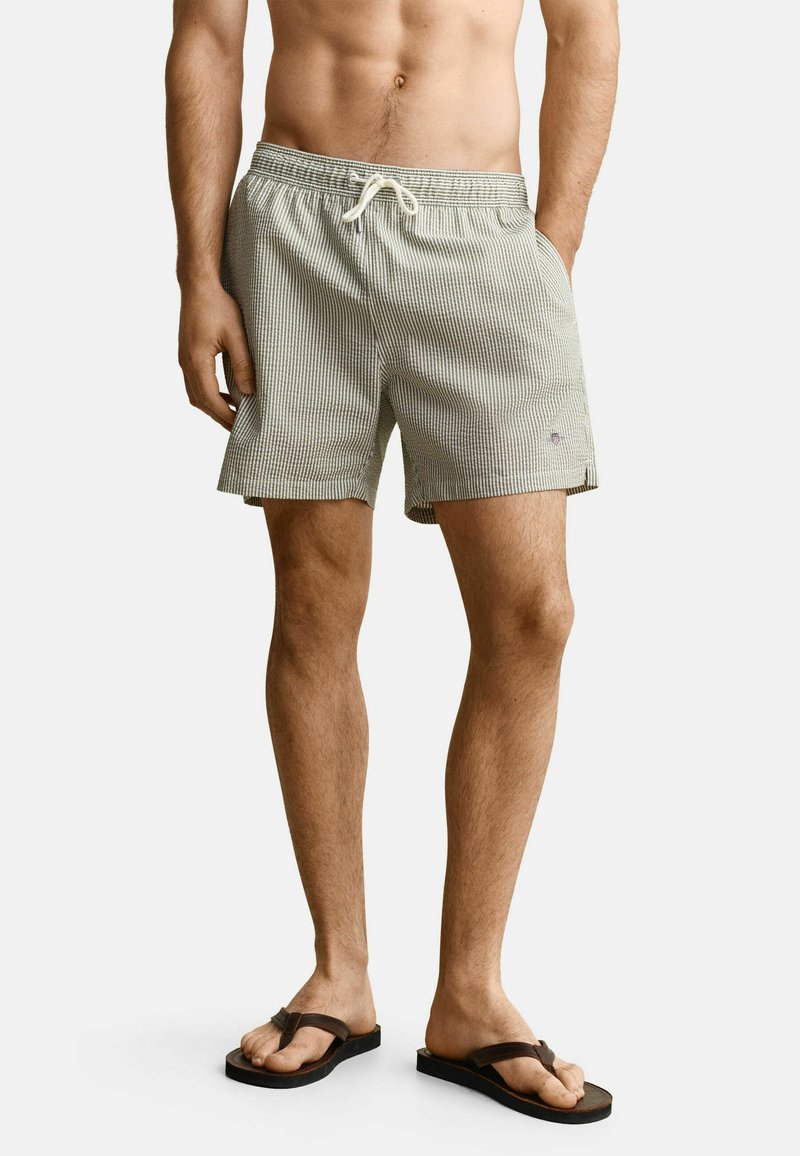 Homme portant un short de bain à rayures gris clair avec cordon de serrage et tongs marron foncé, debout avec une main dans la poche.