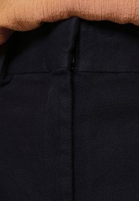 Gros plan sur la ceinture d'un pantalon en tissu noir avec une fermeture éclair latérale cachée, associé à un haut en maille orange.
