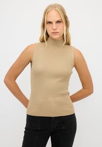 Top senza maniche a coste beige con collo alto e sottili punteggiature texturizzate. Abbinato a jeans scuri, esibendo una silhouette aderente.