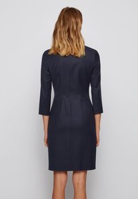 Mörk marinblå, figursydd klänning med knälång nederkant, trekvartslånga ärmar och en slät textur. Har en ren silhuett och subtila sömmar.